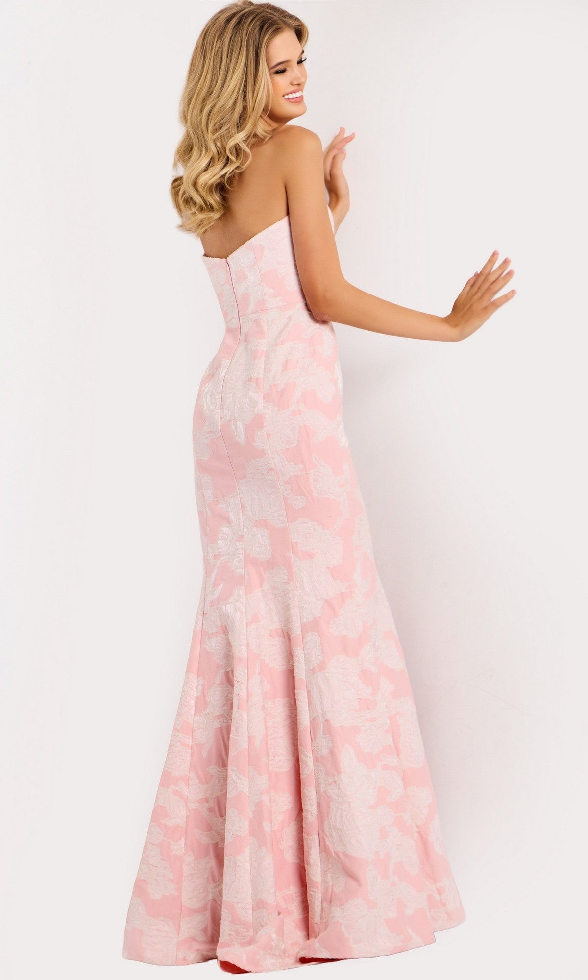 A woman models the Jovani 48260—a strapless long pink prom dress with a delicate floral print—standing against a plain white background, smiling over her shoulder.